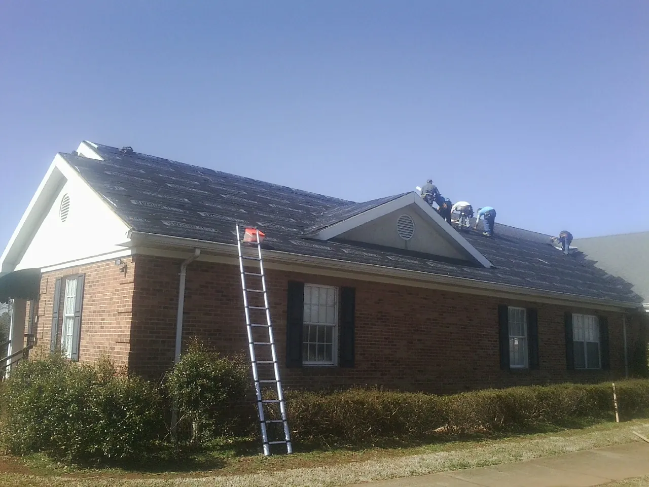 Crew tearing shingles off a brick ranch roof with a ladder set — Emergency Roof Repair work by our crew in Ernul