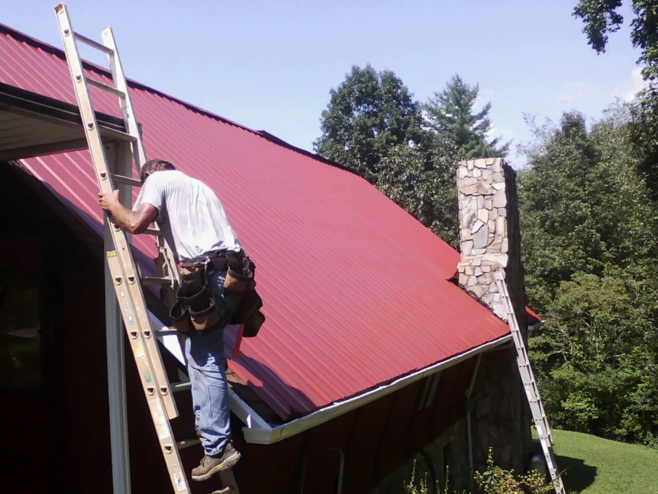 Roofer on a ladder beside a red metal roof and stone chimney — Soffit & Fascia Repair by Roofing Brothers in Ernul